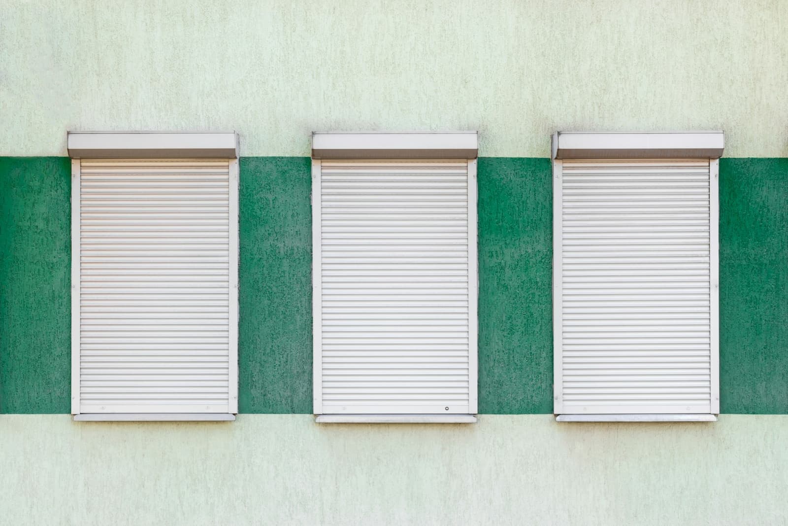Three windows with their roller shutters down on a green and white wall